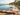 An infinity pool overlooking a tropical ocean resort scene, with stone deck flooring, lounge chairs, palm trees, and buildings in the background.