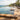 An infinity pool overlooking a tropical ocean resort scene, with stone deck flooring, lounge chairs, palm trees, and buildings in the background.